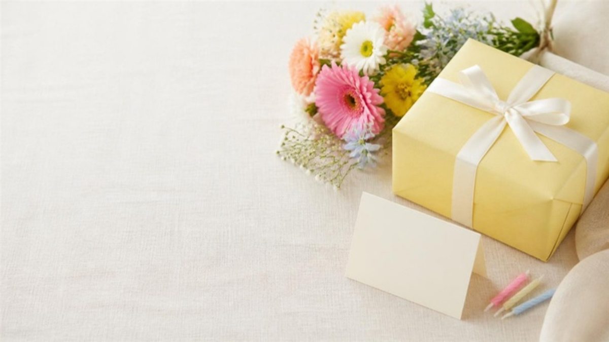 A yellow gift box with a ribbon, a bouquet of gerberas, a blank card and candles